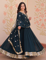 Woman wearing a dark green traditional outfit with gold embroidery against a floral decorated wall.