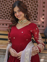 Woman in a red traditional outfit with gold embroidery, standing against an ornate wall.