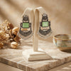 Pair of ornate earrings with green gemstones on a stand, with a blurred background