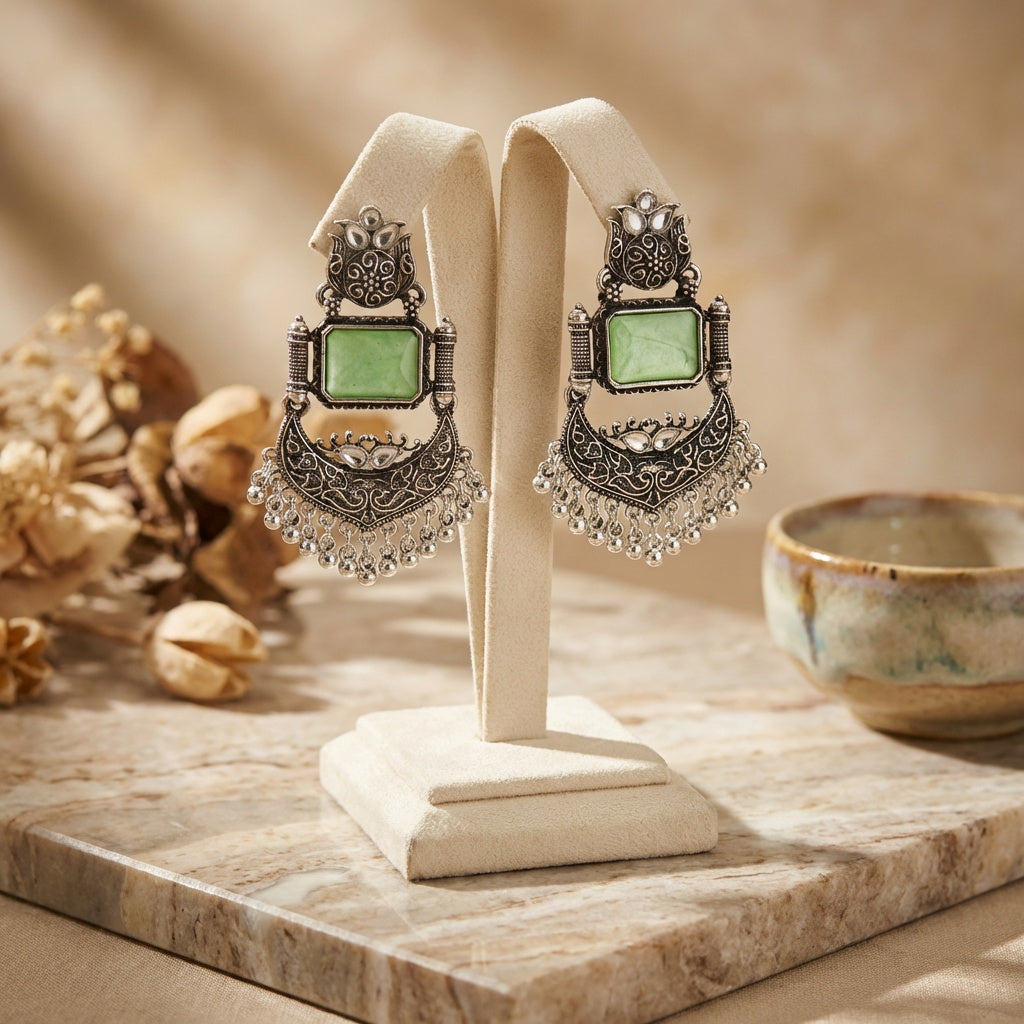 Pair of ornate earrings with green gemstones on a stand, with a blurred background