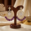 Pair of gold and purple earrings on a wooden stand with a blurred jewelry box in the background.