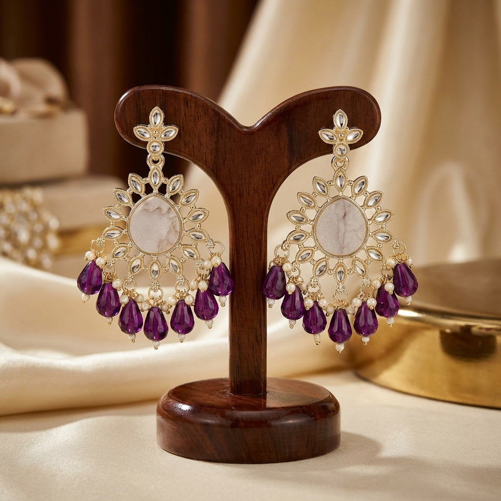 Pair of gold and purple earrings on a wooden stand with a blurred jewelry box in the background.