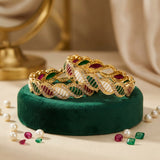 Gold bracelets with gemstones on a green cushion, surrounded by pearls and gemstones.